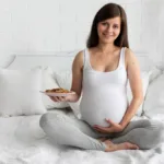 a pregnant woman sitting on a bed holding a plate of cookies